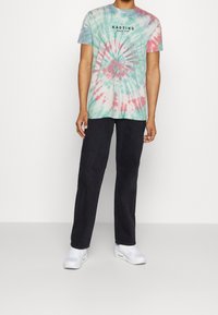 Koszulka tie-dye w miętowym zielonym, różowym i białym kolorze z spiralnym wzorem oraz czarnym napisem "KAOTIKO Since 1999". Czarne spodnie i białe sneakersy.