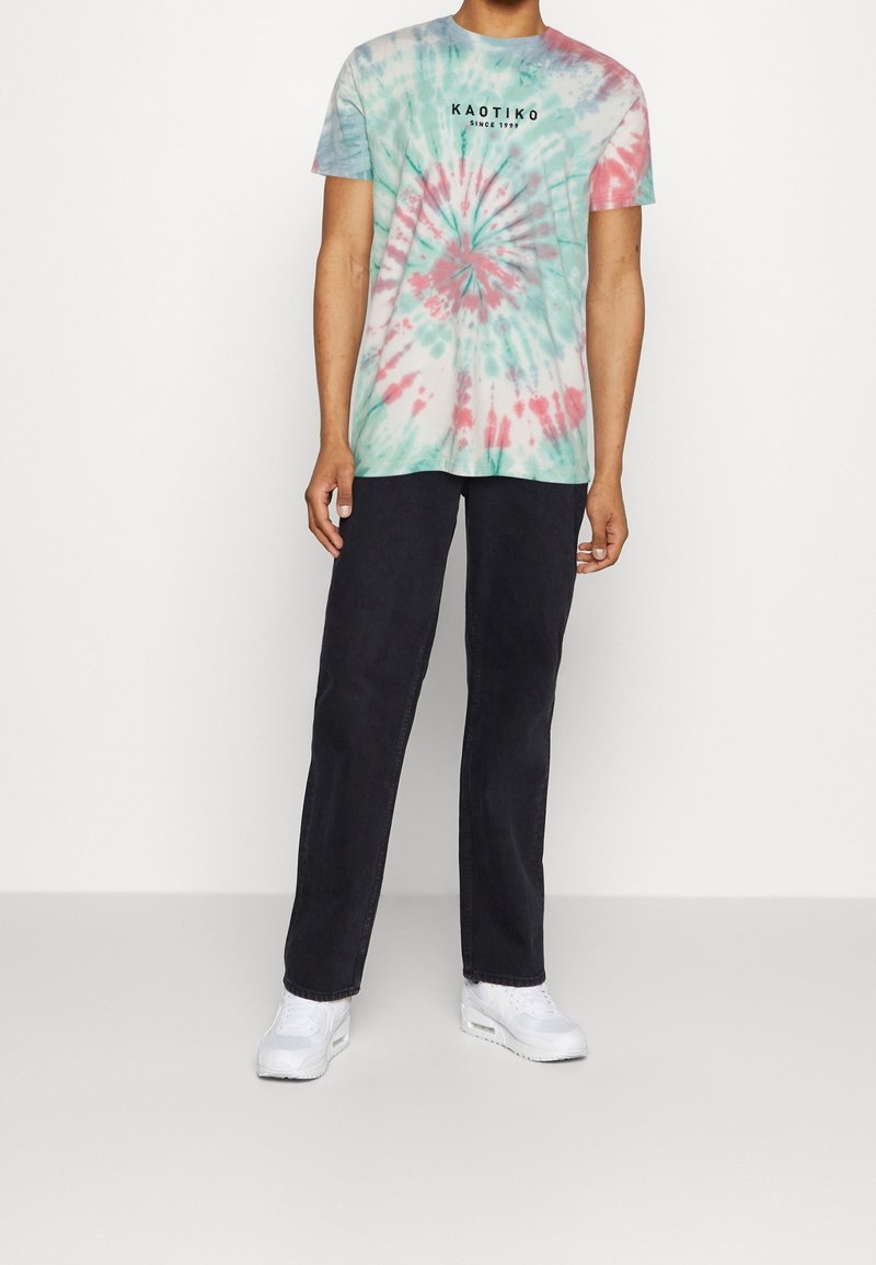 Koszulka tie-dye w miętowym zielonym, różowym i białym kolorze z spiralnym wzorem oraz czarnym napisem "KAOTIKO Since 1999". Czarne spodnie i białe sneakersy.