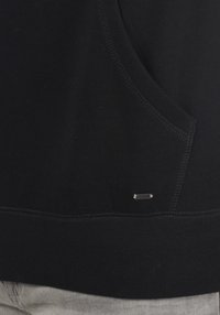 Sudadera negra con textura, que presenta un bolsillo y un pequeño acento de logo plateado cerca del dobladillo. Tela suave con detalles de costura visibles.