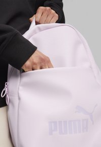 Jasnofioletowy plecak z teksturowanej tkaniny, z kieszenią na zamek i logo PUMA. Projekt charakteryzuje się zaokrąglonym kształtem i eleganckimi akcentami.
