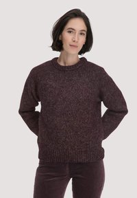 Frau mit kurzem dunklem Haar, die einen dunkelvioletten Strickpullover und eine passende Hose trägt, steht mit den Händen hinter dem Rücken.