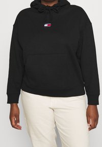 Sudadera negra de tela suave, con un bolsillo canguro en la parte frontal y una capucha ajustable con cordones. Pequeño logo de la marca en el pecho.