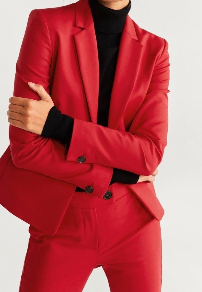 Ensemble blazer et pantalon rouge en tissu lisse ; présente une coupe ajustée, un col roulé noir en dessous et des boutons noirs contrastants sur le blazer.