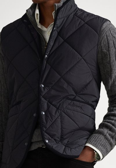 Čierna prešívaná vesta s výplňou, zapínanie na cvoky a bočným vreckom; nosená cez sivý pletený sveter a bielu košeľu s golierom; moderný dizajn.