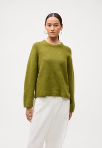 Ženska, ki nosi velike obročaste uhane, olivno zelene puhaste pulover in bele hlače z visokim pasom, stoji pred enobarvno ozadjem.