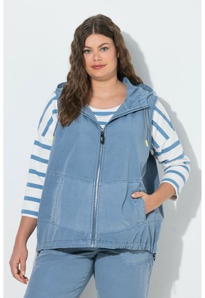 Frau mit welligem braunem Haar trägt eine blaue ärmellose Jacke mit Reißverschluss über einem weiß-blau gestreiften Langarmshirt und passenden blauen Hosen.
