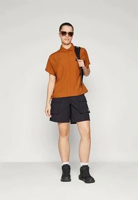 Young person wearing sunglasses, rust-colored short-sleeve shirt, black cargo shorts, black boots, and carrying a black backpack over one shoulder.