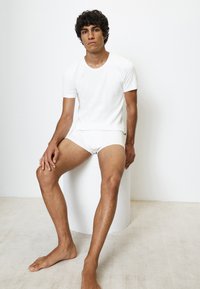 Weißes Baumwoll-T-Shirt mit Rundhalsausschnitt und passenden Unterhosen. Slim-Fit-Design, glatte Textur, getragen von einer sitzenden Person vor einem schlichten Hintergrund.