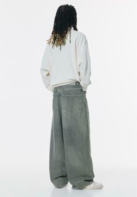 Hanorac supradimensionat de culoare gri deschis, asociat cu blugi verzi din denim, largi și cu talie înaltă. Textură netedă, cu buzunare mari pe spate și o croială lejeră.
