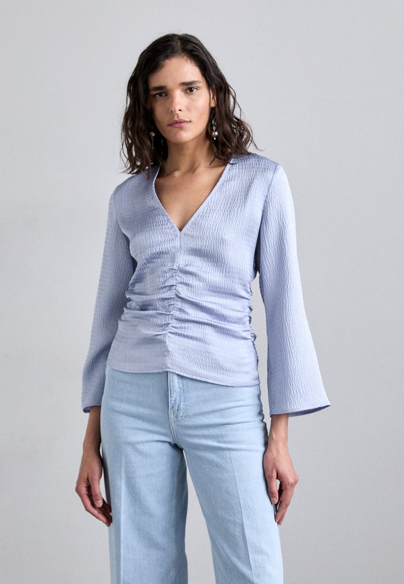 Lys blå, teksturert topp med V-hals, lange bell-ermer og rynkede detaljer i midjen, parret med lys blå jeans.