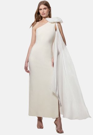 Donna che indossa un abito crema monospalla con un grande dettaglio in tessuto bianco drappeggiato sulla spalla, abbinato a sandali dorati con tacco alto.
