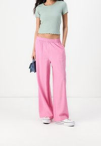 Kvinna i ljusgrön kortärmad ribbad t-shirt, vida rosa byxor, vita sneakers, håller en liten blå handväska mot vit bakgrund.