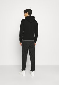 Felpa nera con cappuccio e cordino, polsini e orlo a coste. Abbinata a pantaloni neri larghi e sneakers bianche, vista da dietro.
