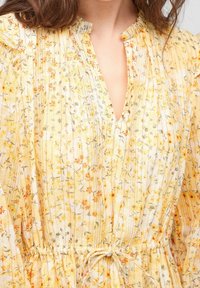 Femme portant une robe jaune à fleurs avec une taille froncée et un col à volants, montrant la partie supérieure de la poitrine et la zone du cou.