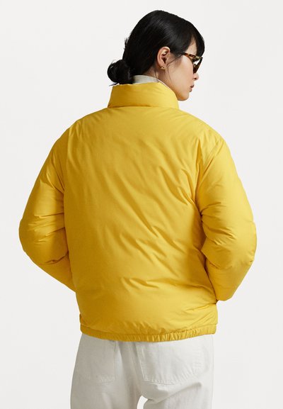 Geel pufferjack met een hoge kraag, gladde textuur, elastische zoom en zijzakken, gedragen over witte broek.