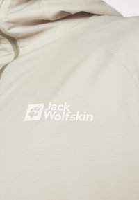 Lys beige jakke med en glat, tekstureret overflade. Har et hvidt logo, der står "Jack Wolfskin", samt et poteaftryk som accent.