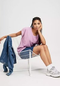 Jasnofioletowa, oversize t-shirt, dżinsowe szorty, białe sneakersy, siedząca na białym drucianym krześle z niebieską dżinsową kurtką zarzuconą na oparcie.