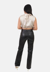 Donna con lunghi capelli scuri, indossa un top beige in satin senza maniche con un fiocco sul retro, pantaloni in pelle nera e scarpe nere con tacco alto, rivolta di spalle.