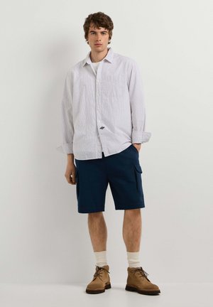 Jonge man met een licht gestreept overhemd, marineblauwe cargoshorts, witte sokken en bruine veterschoenen staand tegen een effen achtergrond.
