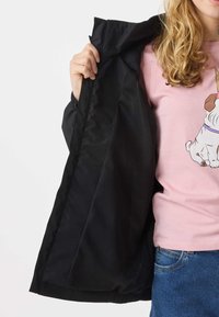 Person with curly blonde hair shows inside of open black jacket, wearing pink t-shirt with dog graphic and blue jeans.