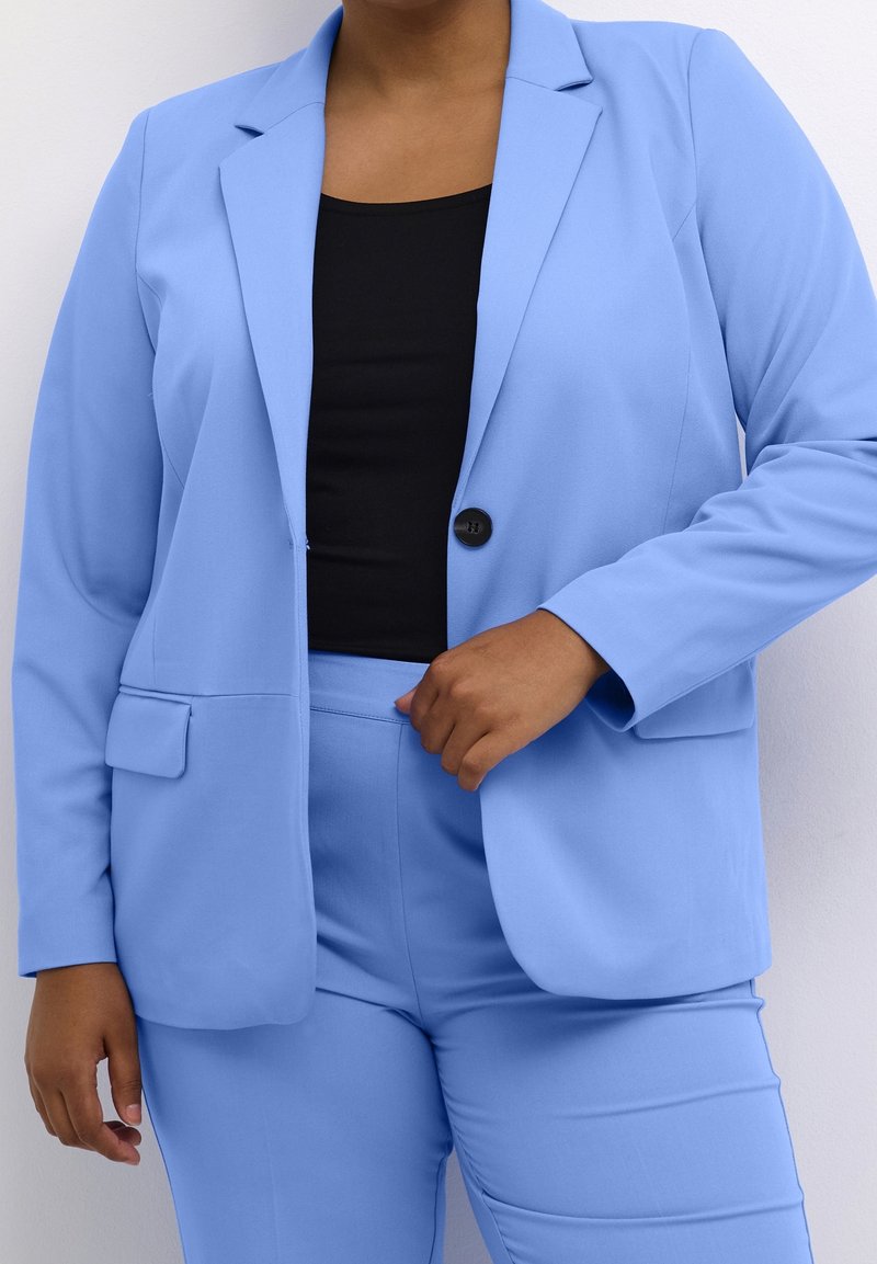 Personne portant un blazer bleu clair et un pantalon assorti avec une chemise noire en dessous, ajustant le blazer près du bouton d'une main.