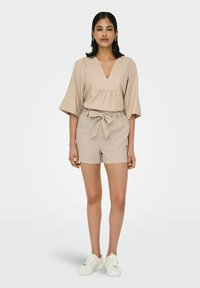 Beige, locker sitzendes V-Ausschnitt-Oberteil mit kurzen Ärmeln, kombiniert mit passendem hoch-tailliertem Shorts und weißen Sneakers. Strukturierter Stoff mit Gürteldetail.