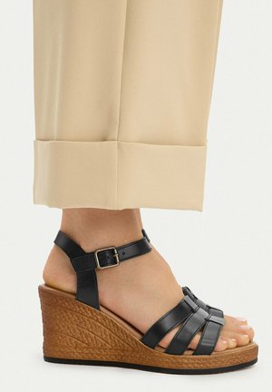 Pie con pantalones beige y sandalia cuña de cuero negro con hebilla en el tobillo y tacón marrón tejido, mostrado de perfil.