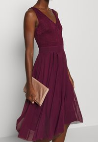 Burgundské šaty s krajkovým korzetem, výstřihem do V a plisovanou sukní. Modelka drží jemně růžovou kabelku a předvádí elegantní design outfitu.