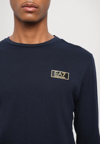 Jūrų laivyno ilgos rankovės marškiniai, pagaminti iš medvilnės, pateikiantys stačiakampį auksinį logotipą krūtinėje. Apvalus iškirpės dizainas ir lygus paviršius.