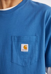 Modrá bavlnená tričko s okrúhlym výstrihom a vreckom na hrudi. Na vrecku je vyšité logo Carhartt v oranžovej a bielej farbe.