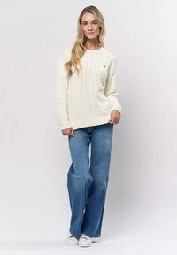 Woman with long blonde hair wearing a cream cable-knit sweater, blue wide-leg jeans, and white sneakers standing against a white background.
