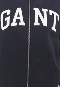Felpa con zip di colore blu navy realizzata in misto cotone, con grandi lettere bianche in rilievo che spellano "GANT" sul davanti, dotata di due tasche laterali.