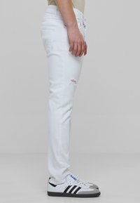 Vaqueros de mezclilla blanca de corte ajustado, con un bolsillo lateral y un ligero desgaste cerca de la rodilla, combinados con zapatillas deportivas en blanco y negro.