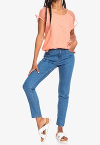 Top corto de coral con acentos de malla con estampado de puntos, combinado con jeans de mezclilla azul ajustados y sandalias blancas de deslizar. Modelo posando de manera casual.
