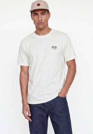 Man wearing beige cap, white R.D.D. printed T-shirt, and dark blue jeans stands with one hand in pocket on white background.