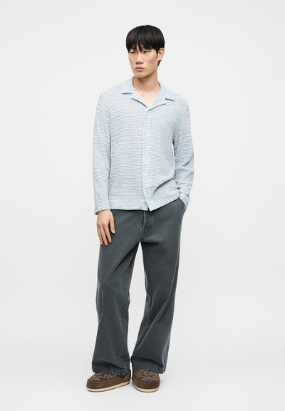 Chemise à rayures bleu clair et blanche avec col, associée à un pantalon large gris foncé et des chaussures marron sans lacets.