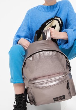 Metalický batoh v růžově zlaté barvě s hladkým povrchem, zaobleným tvarem, přední kapsou na zip a černými popruhy, umístěn vedle osoby v modrém svetru.