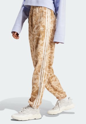 Persona vistiendo pantalones deportivos beige tie-dye de Adidas con rayas blancas y zapatillas blancas, combinados con una camiseta de manga larga color lila claro.