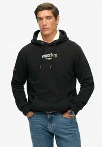 Czarna bluza z kapturem z sznurkami i przednią kieszenią. Nadrukowany logo w kolorze białym. Noszona z niebieskimi dżinsami. Materiał wydaje się miękki i teksturowany.