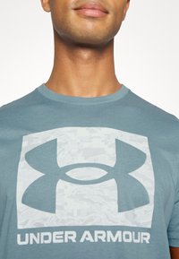 Světle modré sportovní tričko s kulatým výstřihem, zdobené velkým bílým logem Under Armour a jemným maskovacím vzorem v pozadí.