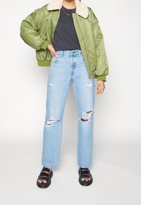 Grön bomberjacka med krämfärgad shearlingkrage, ihop med ljusblå slitna vida jeans och svarta sandalinspirerade skor.