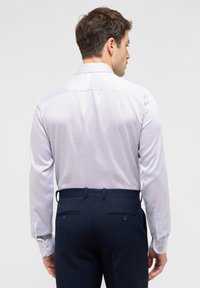 Camicia bianca a maniche lunghe con un motivo geometrico azzurro, abbinata a pantaloni blu scuro. Presenta polsini con bottoni e un colletto classico.