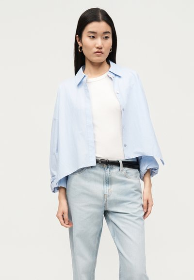 Γυναίκα με ανοιχτό μπλε κοντομάνικο πουκάμισο πάνω από λευκό τοπ, ανοιχτό μπλε τζιν με μαύρη ζώνη και χρυσούς κρίκους στα αυτιά, κοιτάζοντας πλάγια.