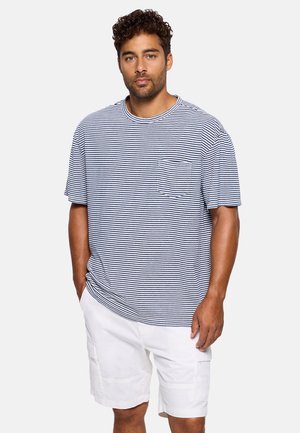 Hombre con cabello rizado que lleva una camisa de manga corta a rayas azules y blancas y pantalones cortos cargo blancos, de pie con las manos en los bolsillos.