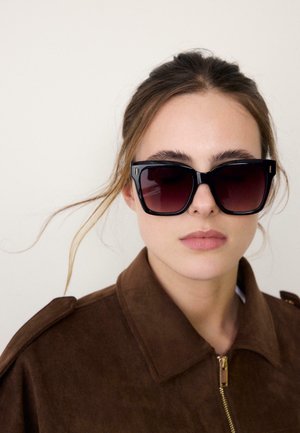 Jeune femme aux cheveux bruns tirés en arrière, portant de grandes lunettes de soleil noires et une veste en daim marron avec une fermeture éclair dorée.