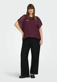 Top in chiffon prugna con maniche larghe, abbinato a pantaloni neri a gamba larga. Il modello indossa scarpe chunky nere e si trova davanti a uno sfondo neutro.