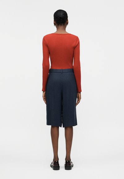 Γυναίκα με σκούρο δέρμα και μαλλιά σε κότσο φοράει κόκκινο μακρυμάνικο τοπ, μπλε σκούρες, πλατιές βερμούδες μέχρι το γόνατο και μαύρα μοκασίνια, στέκεται με την πλάτη.