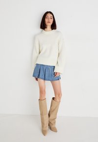 Krem pulover iz pletene tkanine, denim mini krilo z naborki in bež škornji do kolen. Mehak material, sproščen kroj, rebrasti manšeti in detajli z ostrim nosom.