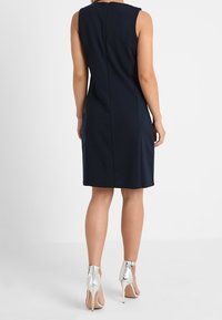 Granatowa sukienka bez rękawów o dopasowanej sylwetce, z zamkiem błyskawicznym z tyłu, gładki materiał, zestawiona z srebrnymi butami na wysokim obcasie.