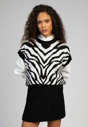 Femme aux cheveux bouclés portant un gilet en tricot à imprimé zèbre noir et blanc sur une chemise blanche et une jupe noire, debout devant un fond gris.